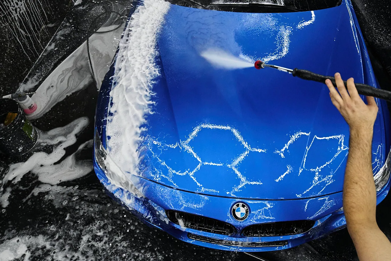 Струя воды сбивает пену с капота синего седана BMW, на лаке остаётся рисунок сот